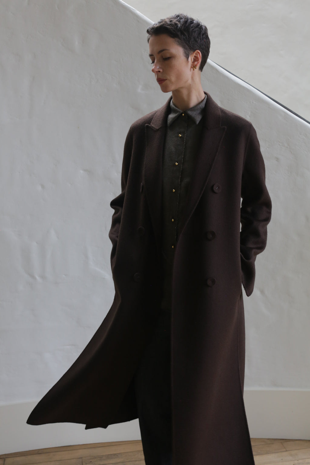 A person with short dark hair, wearing the Doublebreasted Coat | Watermark Brown and a buttoned shirt, stands indoors with hands in pockets and eyes closed against a white textured wall.