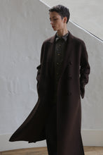 Load image into Gallery viewer, A person with short dark hair, wearing the Doublebreasted Coat | Watermark Brown and a buttoned shirt, stands indoors with hands in pockets and eyes closed against a white textured wall.