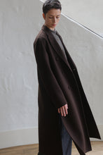 Load image into Gallery viewer, A person with short dark hair stands indoors against a white textured wall, looking down, wearing the Doublebreasted Coat | Watermark Brown over a gray collared shirt and dark pants.