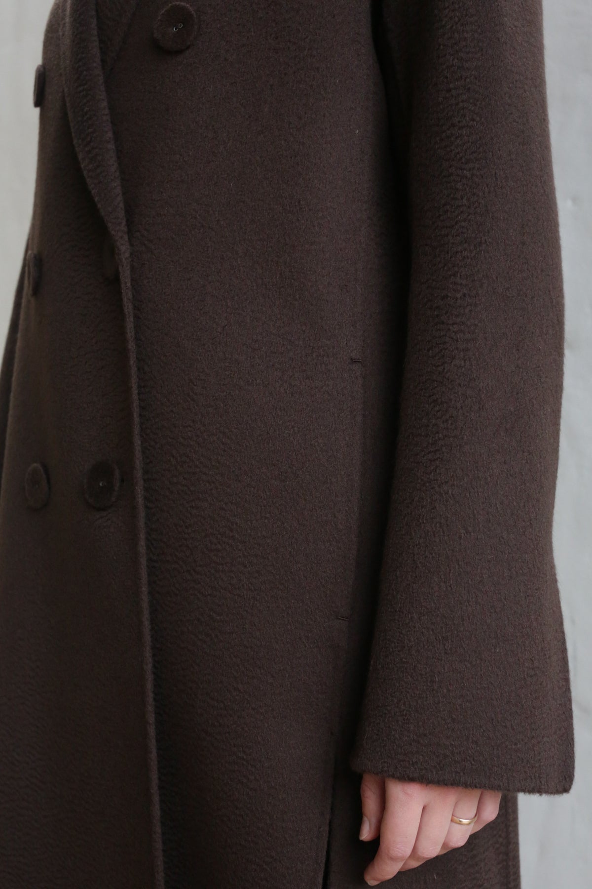 A close-up of a person wearing the Doublebreasted Coat | Watermark Brown shows large lapels and buttons, with only part of the torso and one arm visible, the hand partly tucked into the side seam pocket.