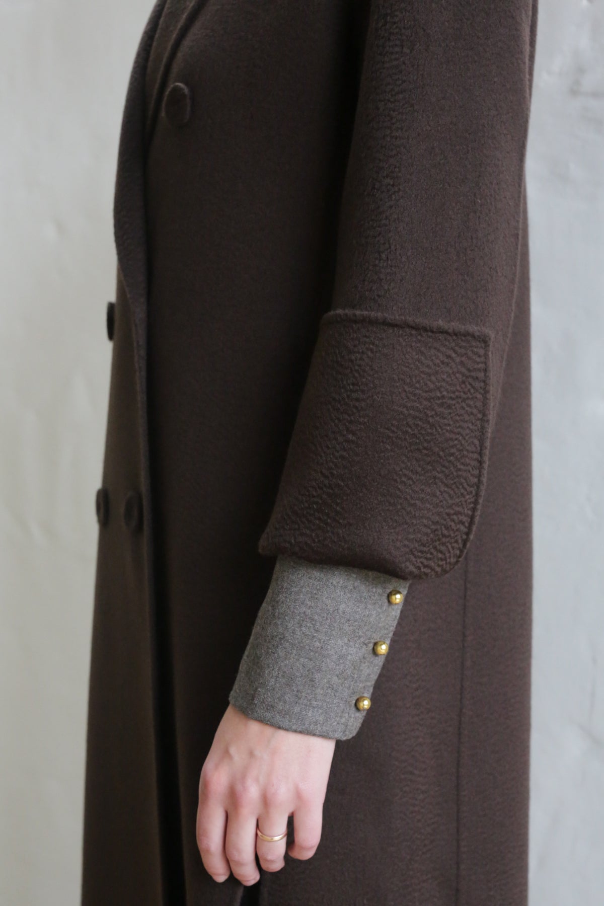 A person wears the Doublebreasted Coat | Watermark Brown, a dark brown textured coat with wide cuffs, over a gray sleeve with three gold buttons. Their hand, adorned with a gold ring, rests at their side against a light background.