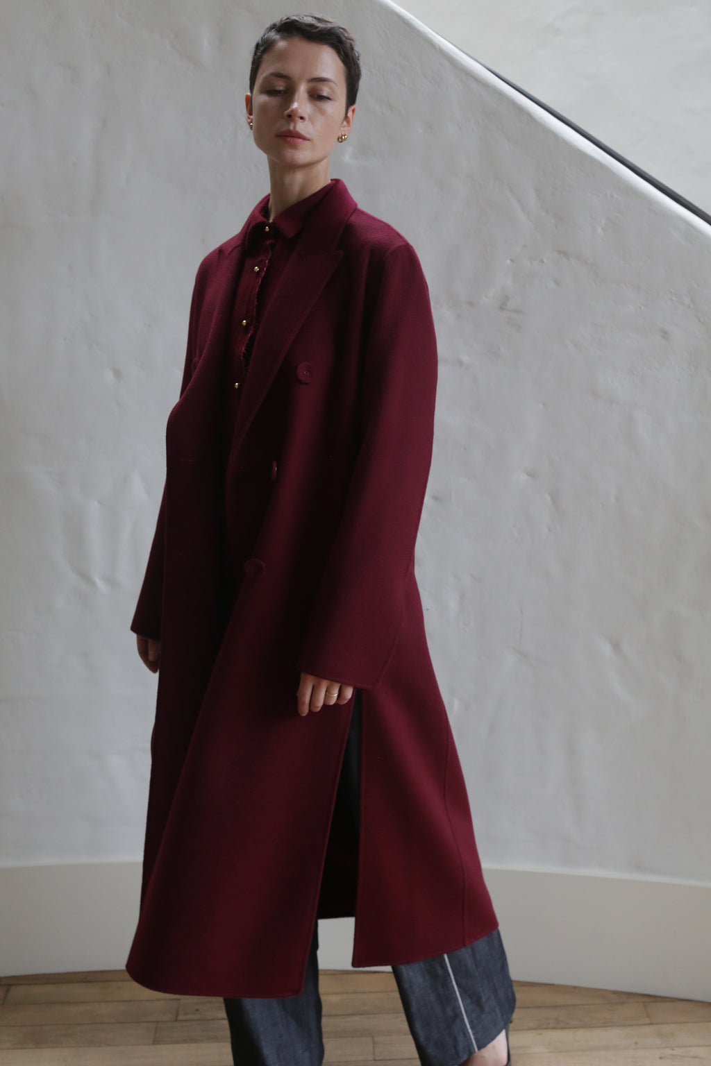 A person with short dark hair wears the Doublebreasted Coat | Watermark Burgundy over dark pants, standing indoors against a white textured wall and wooden floor. The calm expression and soft lighting enhance the elegant look.