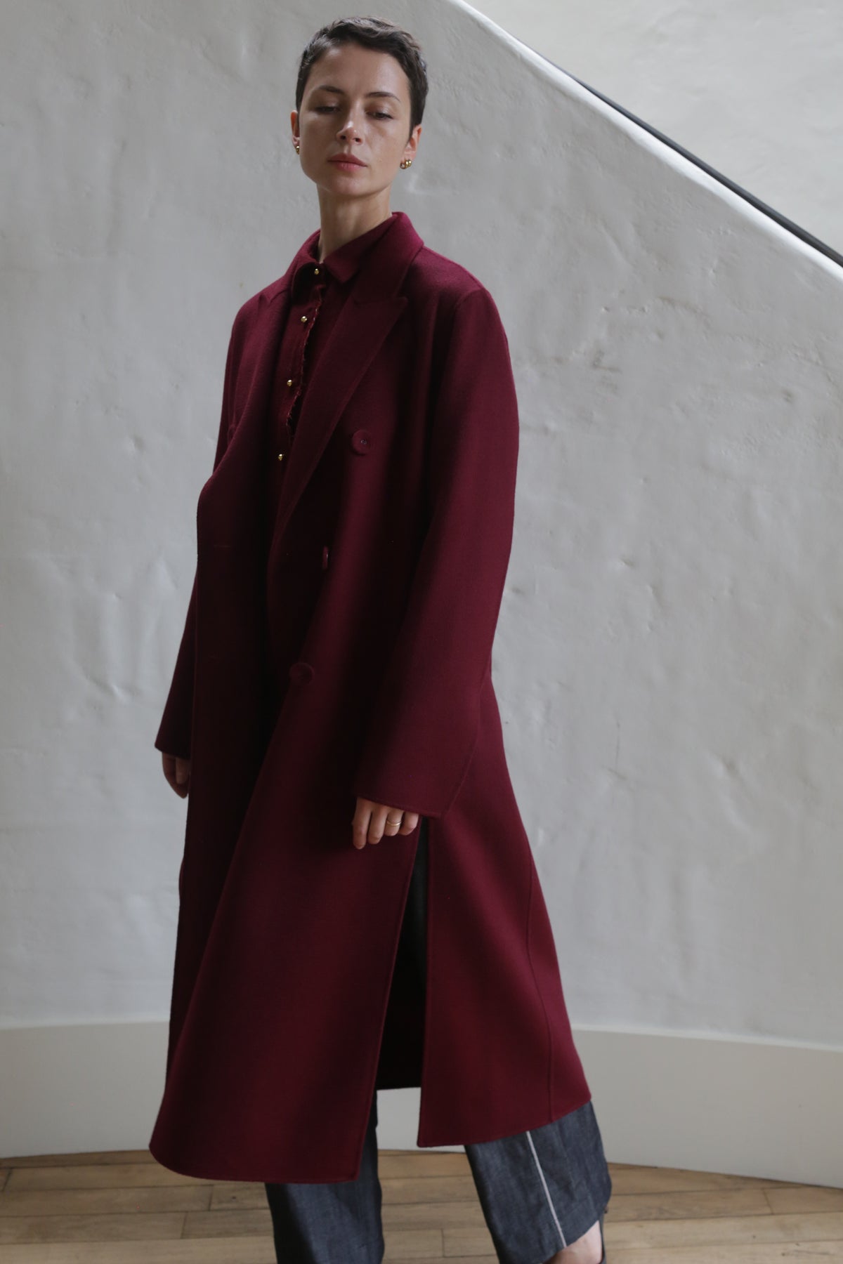 A person with short dark hair wears the Doublebreasted Coat | Watermark Burgundy over dark pants, standing indoors against a white textured wall and wooden floor. The calm expression and soft lighting enhance the elegant look.
