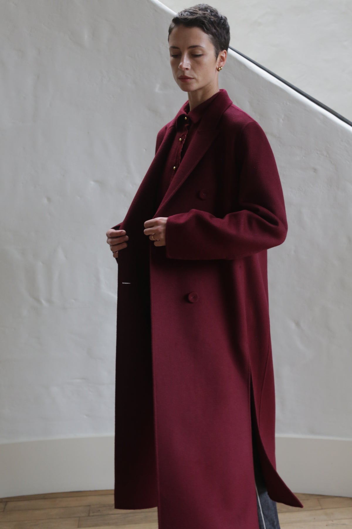 A person wearing the Doublebreasted Coat | Watermark Burgundy stands indoors against a white textured wall, holding the coat open slightly with both hands.