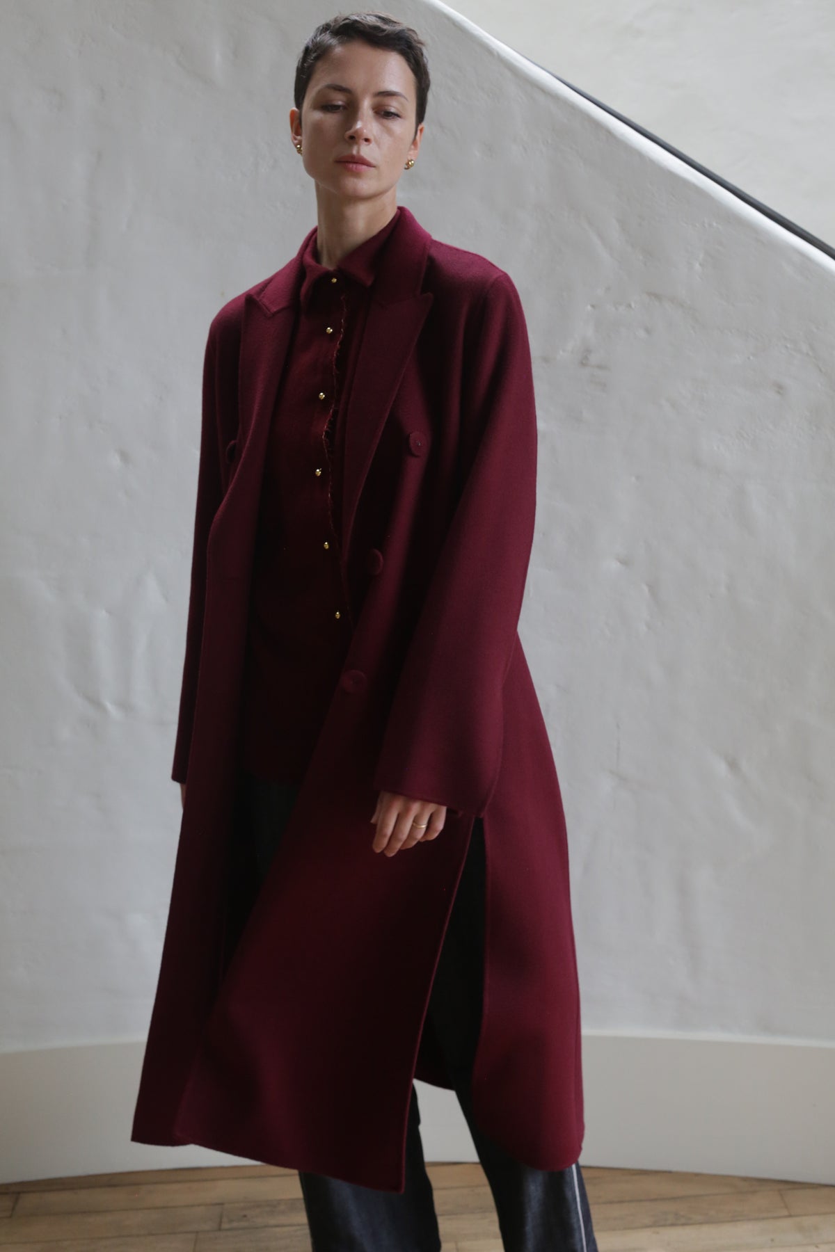 A person with short dark hair wears the Doublebreasted Coat | Watermark Burgundy over a matching shirt and dark pants, standing indoors against a white textured wall and wooden floor.