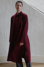 Load image into Gallery viewer, A person with short dark hair wears the Doublebreasted Coat | Watermark Burgundy over a matching shirt and dark pants, standing indoors against a white textured wall and wooden floor.