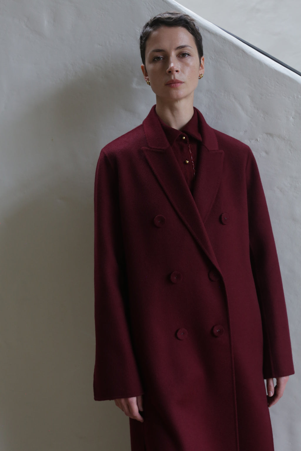 A person with short hair stands against a light wall, wearing the Doublebreasted Coat | Watermark Burgundy over a matching shirt.