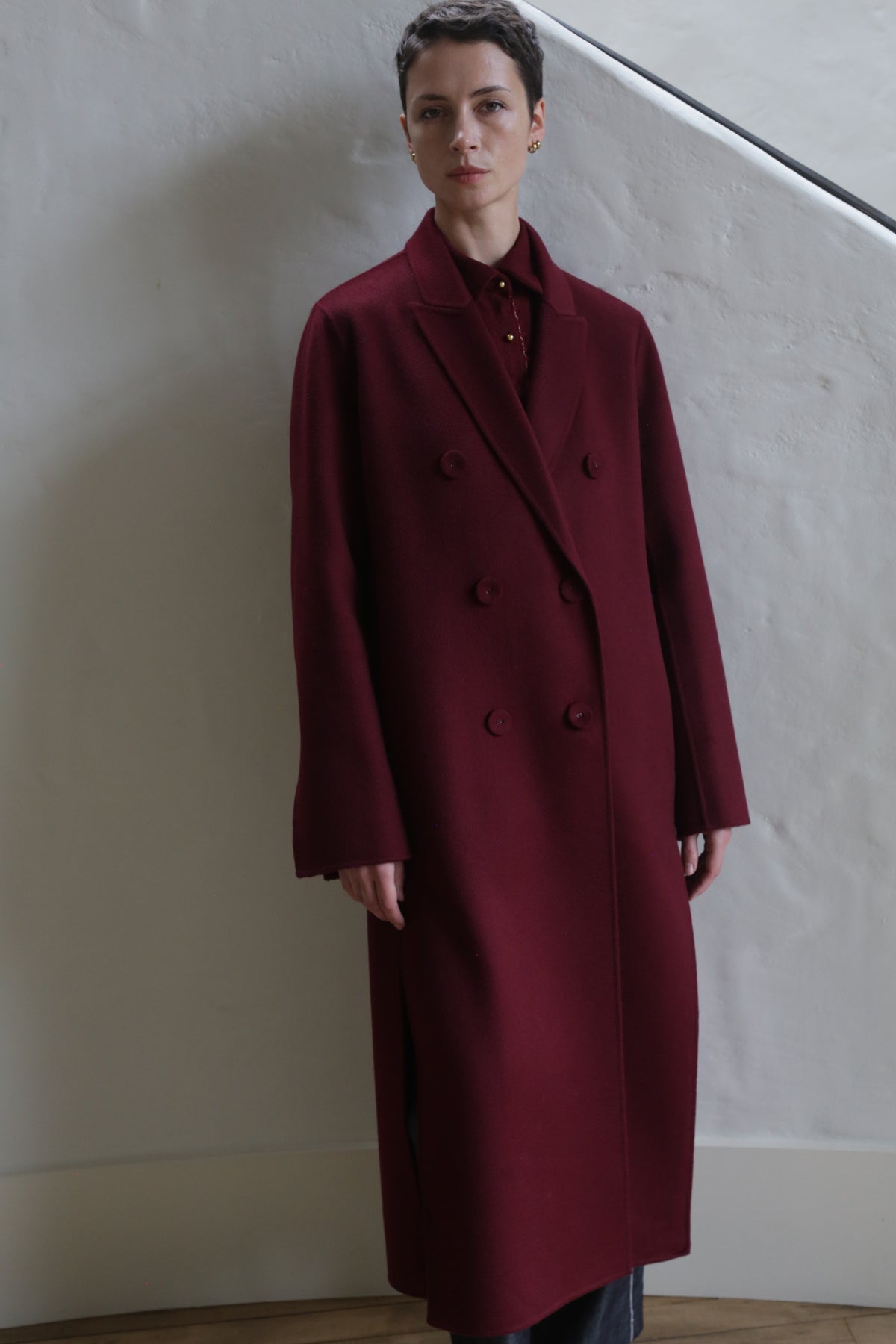 A person with short dark hair is wearing the Doublebreasted Coat | Watermark Burgundy and standing indoors against a light-colored textured wall.