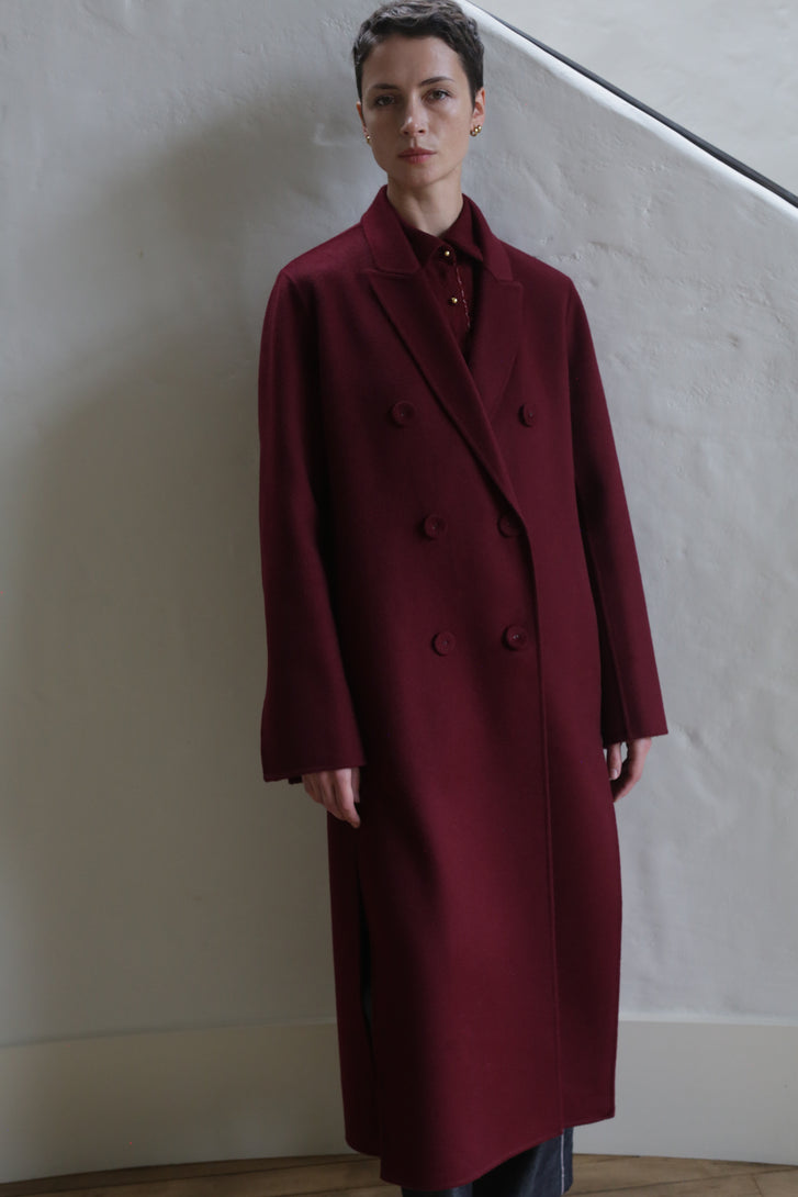 A person with short dark hair is wearing the Doublebreasted Coat | Watermark Burgundy and standing indoors against a light-colored textured wall.