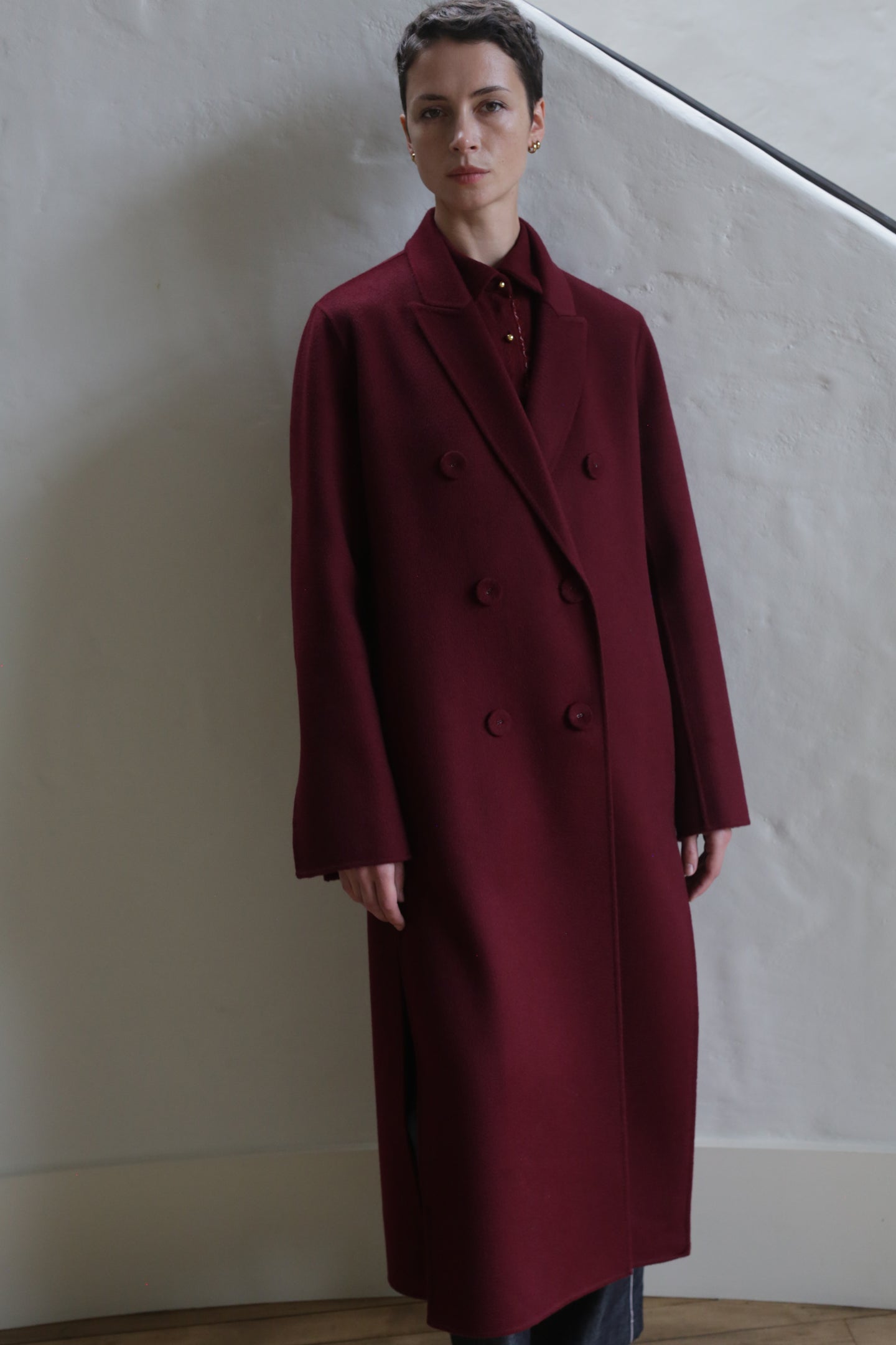 A person with short dark hair is wearing the Doublebreasted Coat | Watermark Burgundy and standing indoors against a light-colored textured wall.
