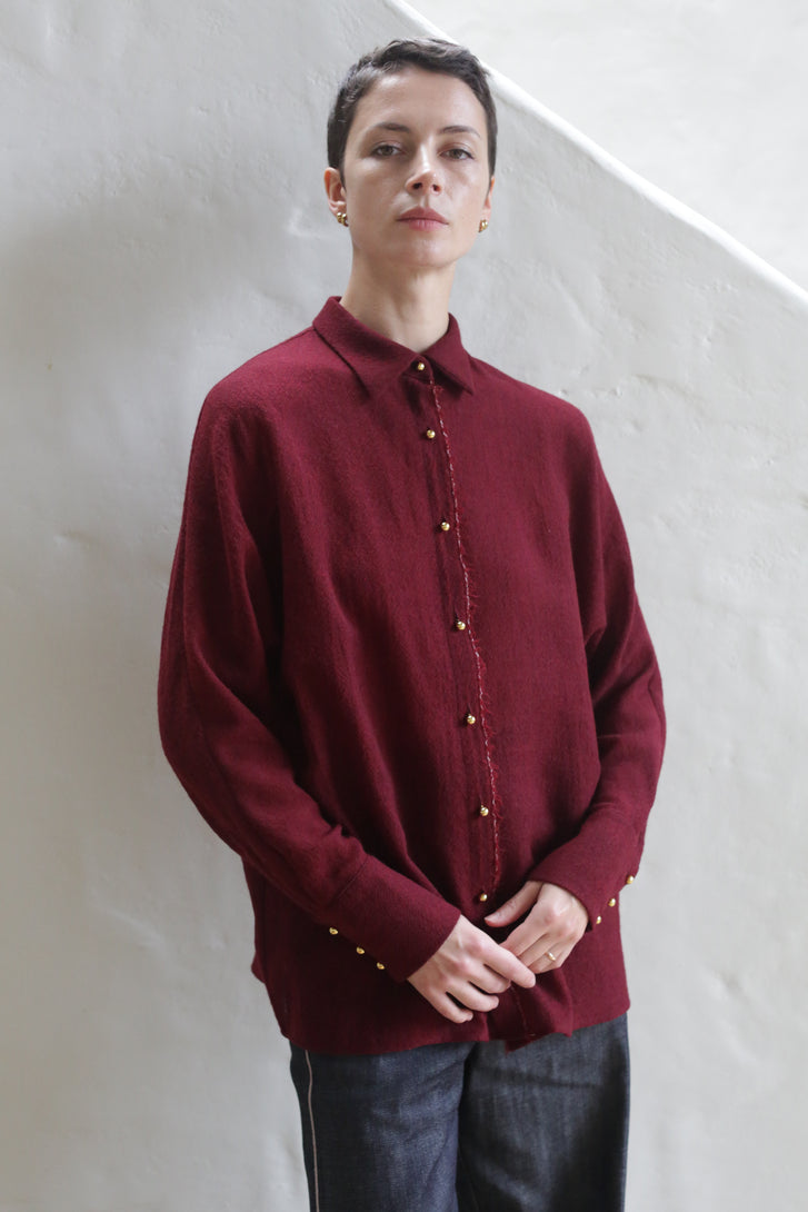 A person with short dark hair wears the Work Shirt | Burgundy and dark pants, standing against an off-white textured wall while looking confidently at the camera.