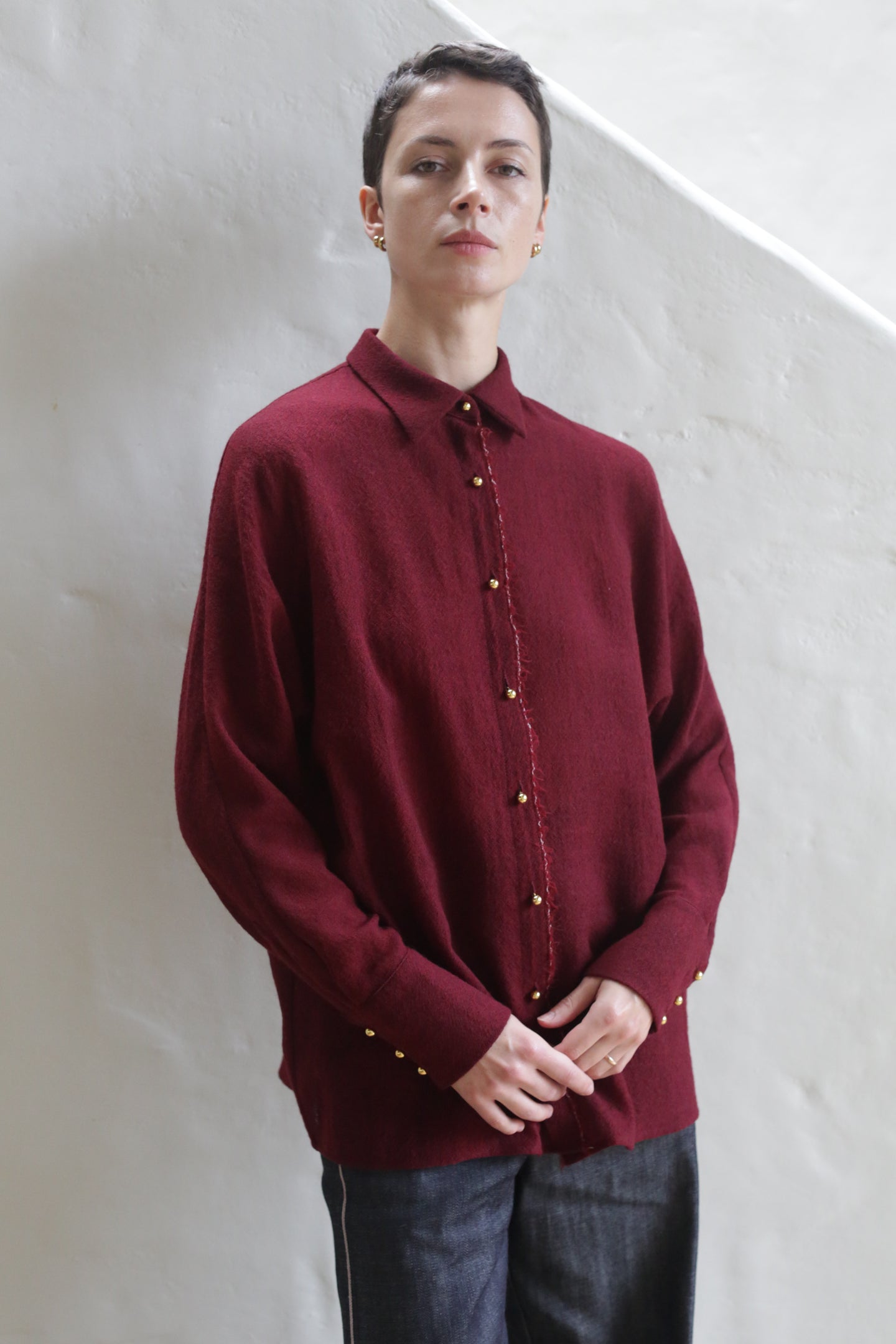 A person with short dark hair wears the Work Shirt | Burgundy and dark pants, standing against an off-white textured wall while looking confidently at the camera.