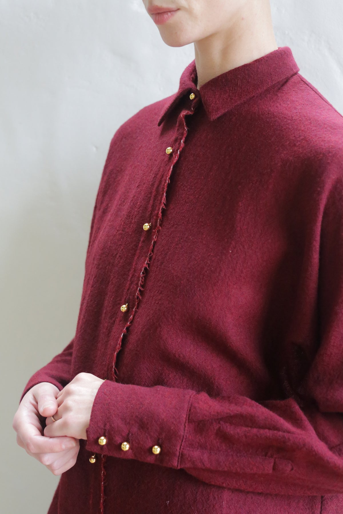 Someone wears the Work Shirt | Burgundy, a long-sleeved shirt with gold buttons on the front and cuffs, standing against a light background. Their face is partially out of frame.