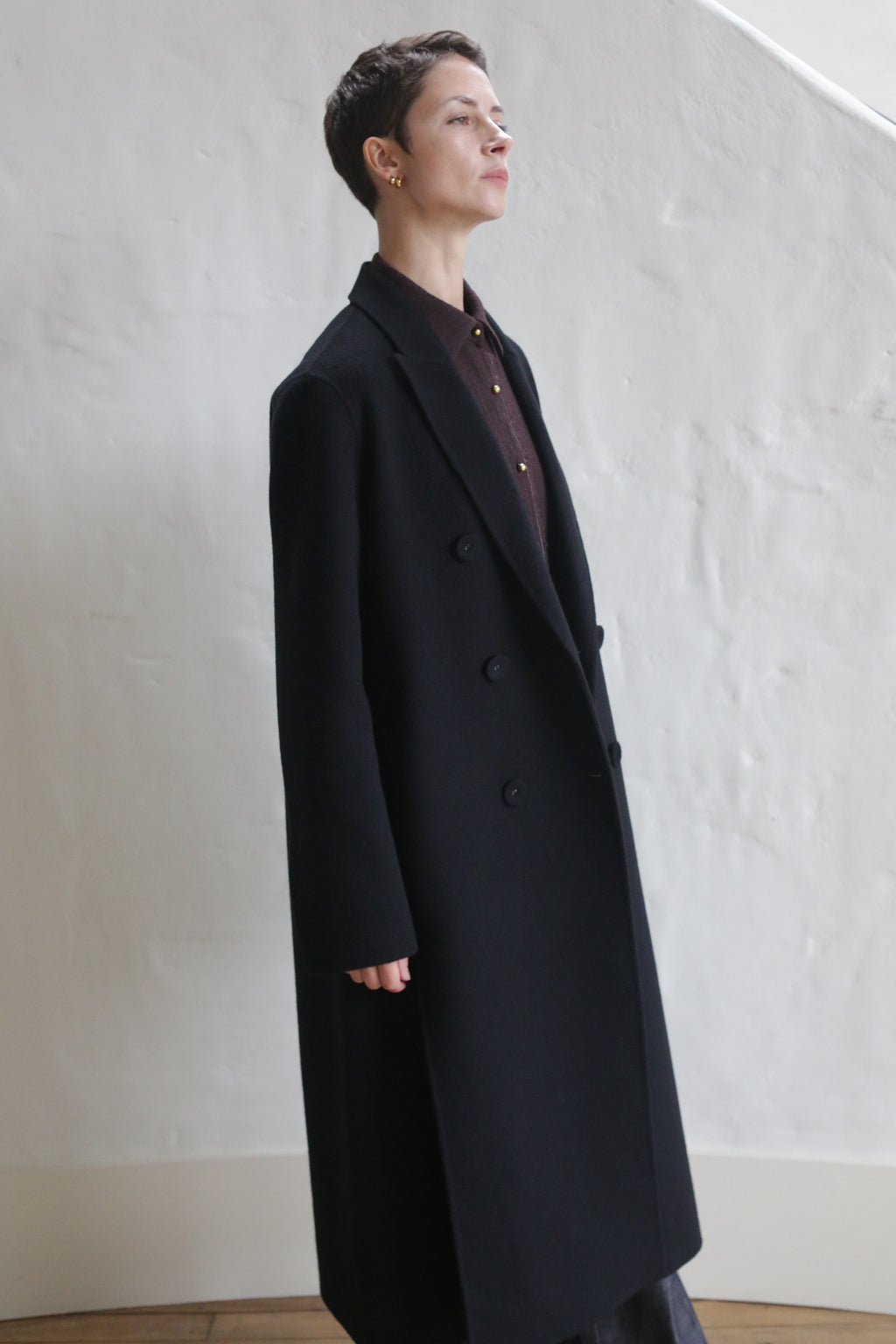 A person with short hair wears the Doublebreasted Coat | Watermark Black over a dark shirt, standing indoors against a white textured wall.