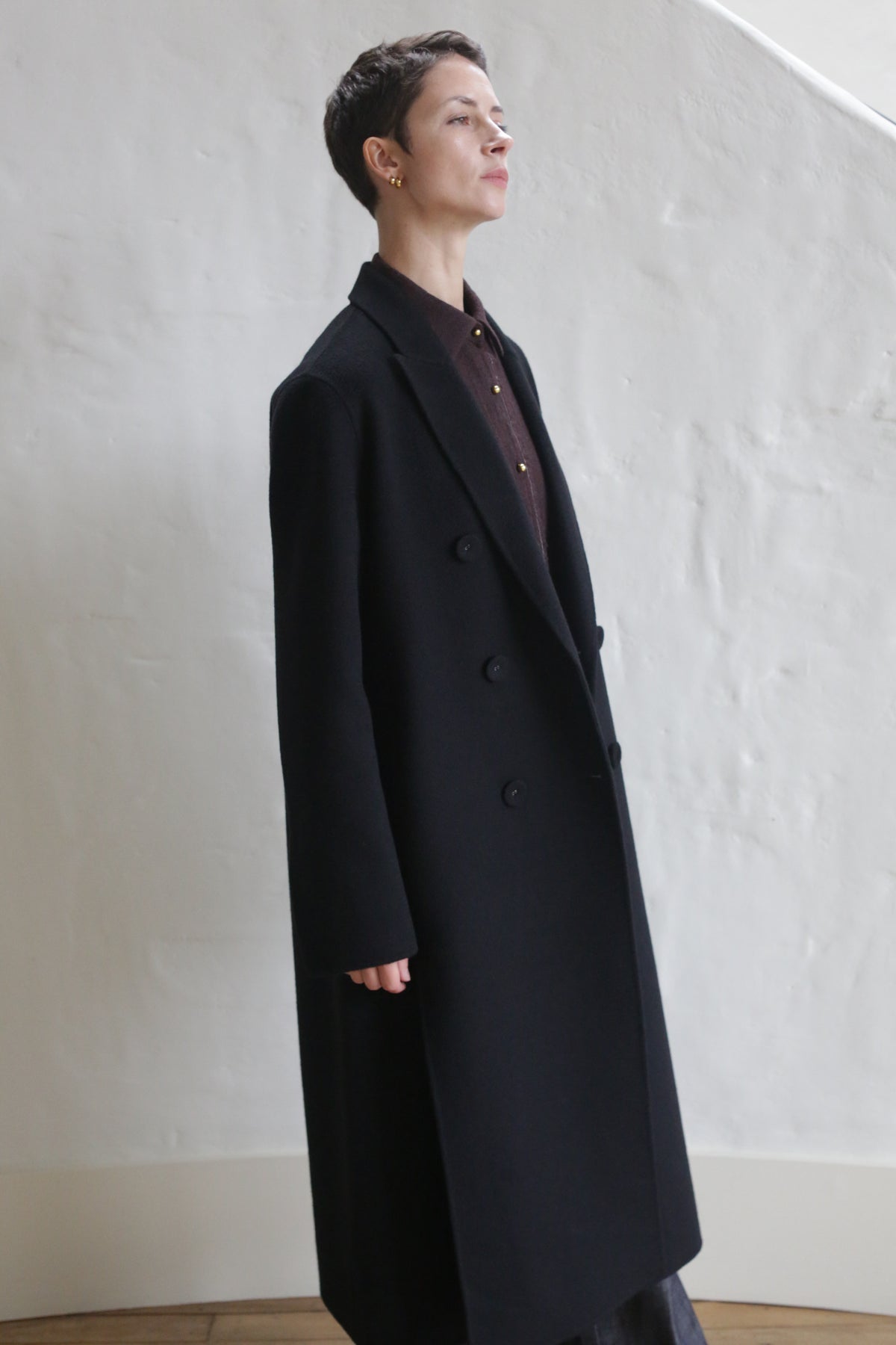 A person with short hair wears the Doublebreasted Coat | Watermark Black over a dark shirt, standing indoors against a white textured wall.