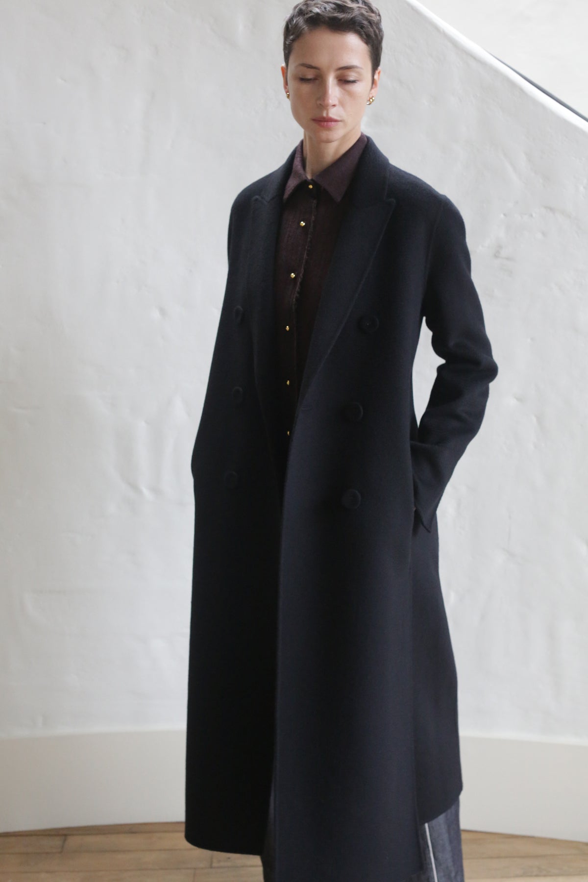A person with short hair stands indoors against a white textured wall, wearing the Doublebreasted Coat | Watermark Black over a dark button-up shirt, hands in pockets and eyes slightly downcast.