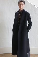 Load image into Gallery viewer, A person with short hair stands indoors against a white textured wall, wearing the Doublebreasted Coat | Watermark Black over a dark button-up shirt, hands in pockets and eyes slightly downcast.