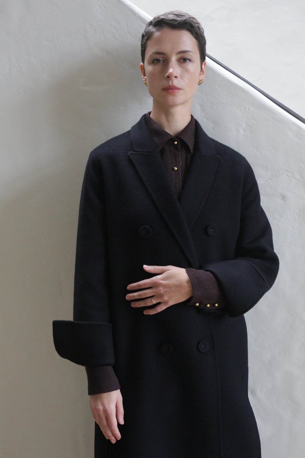 A person with short hair wears the Doublebreasted Coat | Watermark Black over a dark shirt indoors, standing against a pale wall with one arm bent and resting lightly on their midsection.