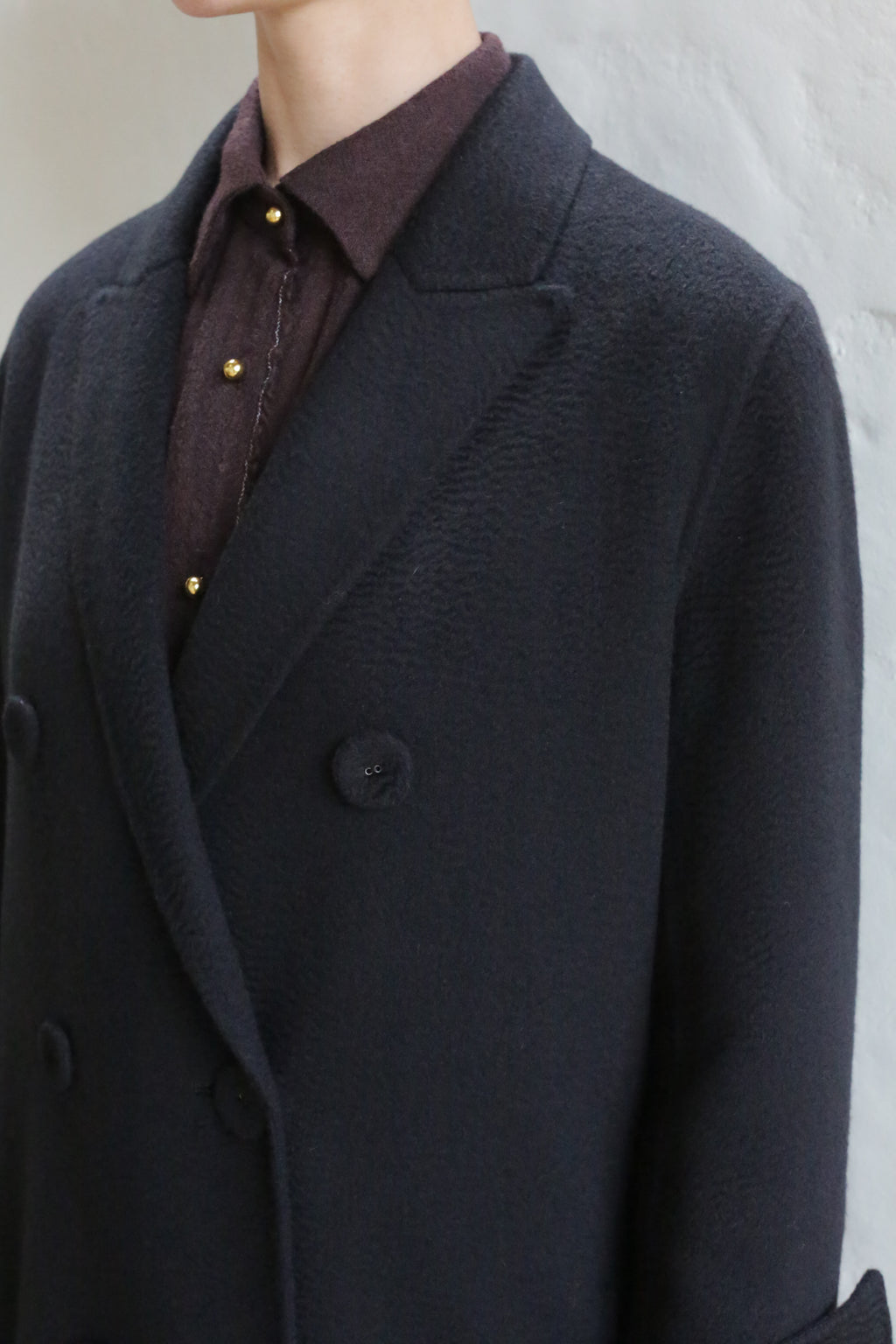 A person wearing the Doublebreasted Coat | Watermark Black over a brown collared shirt with gold buttons, standing against a light-colored wall with their head turned slightly to the side.