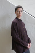 A person with short dark hair, wearing the Work Shirt in Aubergine, stands indoors against a light-colored textured wall and diagonal black railing, looking calmly at the camera.