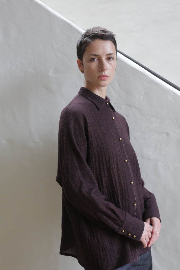 A person with short dark hair, wearing the Work Shirt in Aubergine, stands indoors against a light-colored textured wall and diagonal black railing, looking calmly at the camera.
