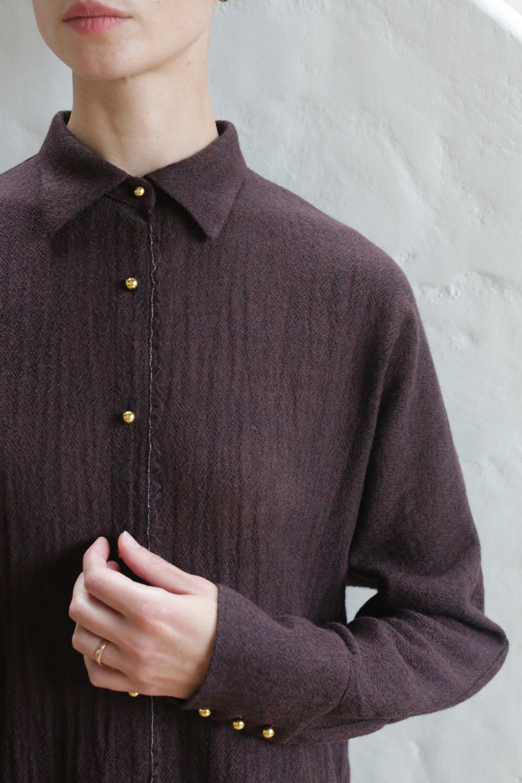 A person wearing the Work Shirt | Aubergine stands against a white textured wall, touching the front of the shirt. Their face is partially out of frame.