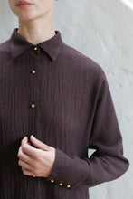 Load image into Gallery viewer, A person wearing the Work Shirt | Aubergine stands against a white textured wall, touching the front of the shirt. Their face is partially out of frame.