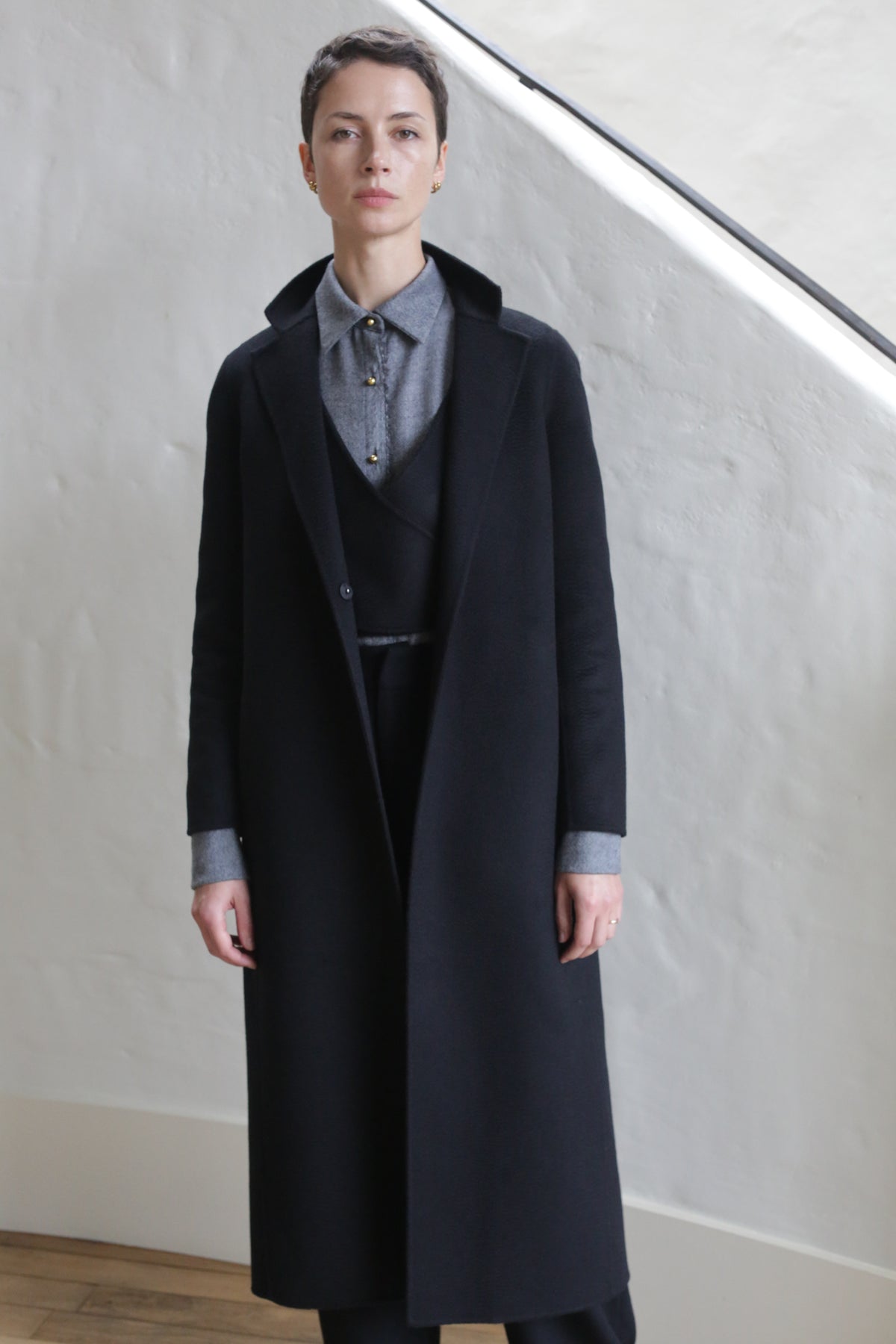 A person with short hair stands indoors, wearing the X-Scarf Coat | Watermark Black over a gray collared shirt and dark trousers, posing against a light-colored textured wall.
