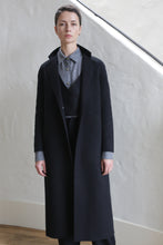 Load image into Gallery viewer, A person with short hair stands indoors, wearing the X-Scarf Coat | Watermark Black over a gray collared shirt and dark trousers, posing against a light-colored textured wall.
