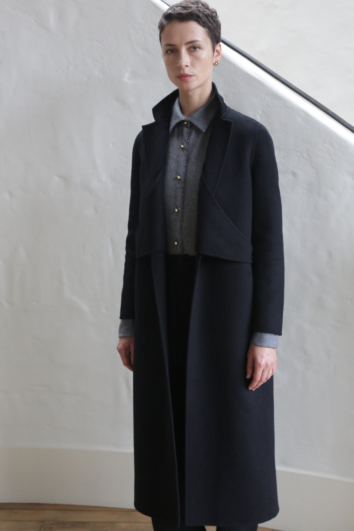 A person with short hair stands indoors against a white textured wall, wearing the X-Scarf Coat | Watermark Black over a gray collared shirt with gold buttons. They appear calm, hands relaxed at their sides.