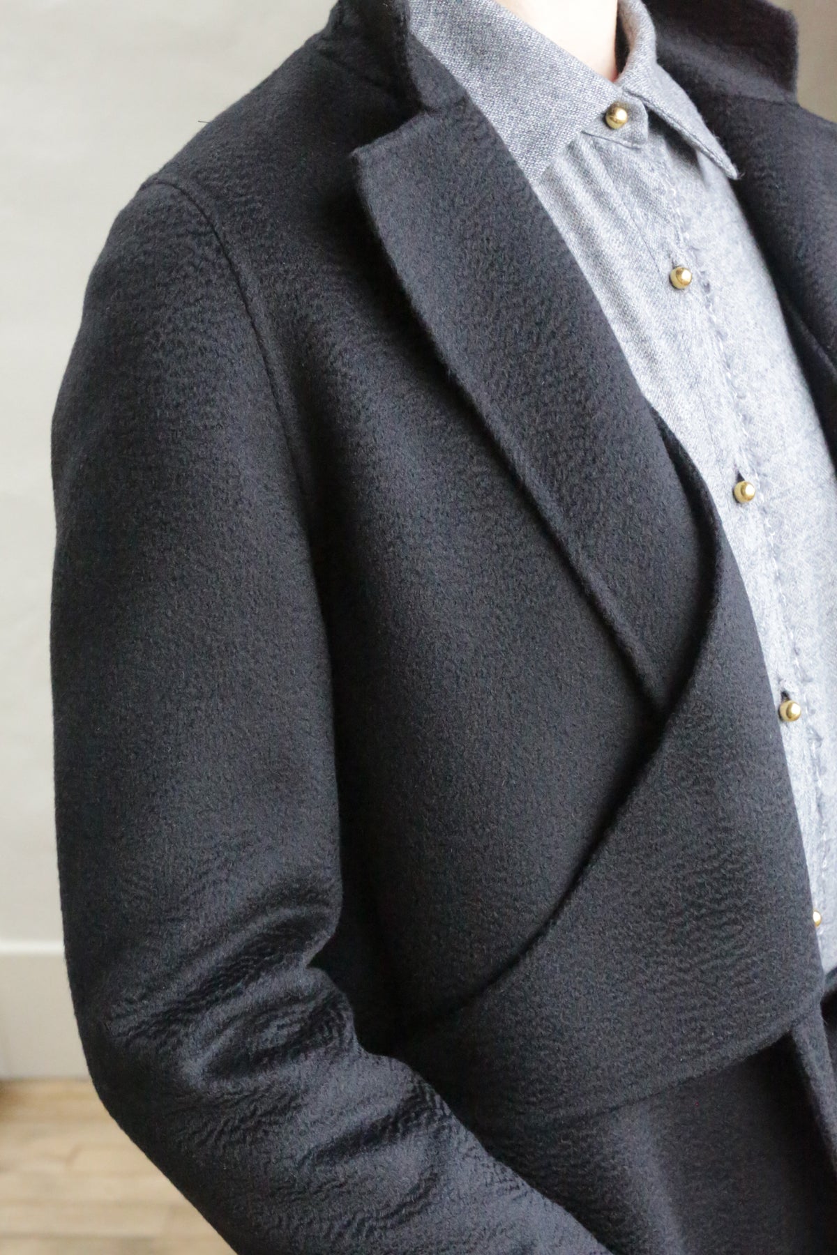A person wearing the X-Scarf Coat in Watermark Black over a light gray button-up shirt with gold buttons, standing indoors against a light-colored wall.