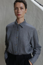 Load image into Gallery viewer, A person with short dark hair wears the Work Shirt | Grey, featuring gold buttons, and black pants while standing with hands in pockets against a light-colored textured wall.