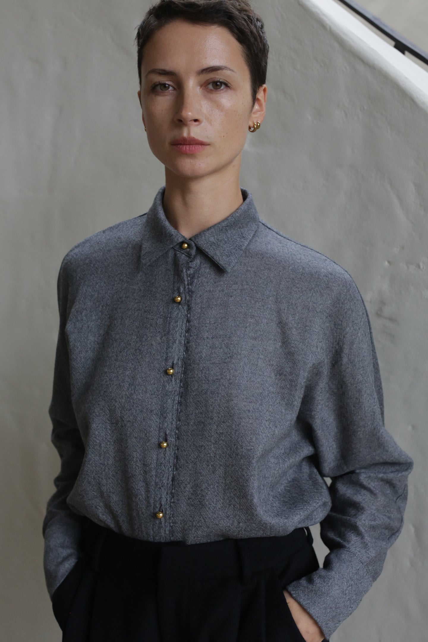 A person with short dark hair wears the Work Shirt | Grey, featuring gold buttons, and black pants while standing with hands in pockets against a light-colored textured wall.