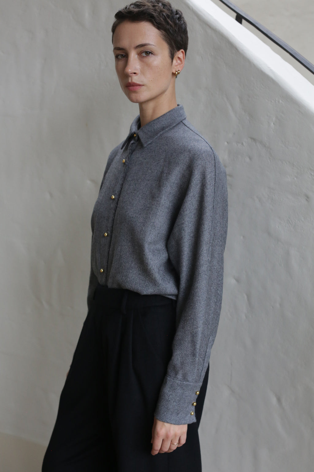 A person with short hair, dressed in the Work Shirt | Grey and black pants, stands beside a light-colored wall, looking calmly at the camera.