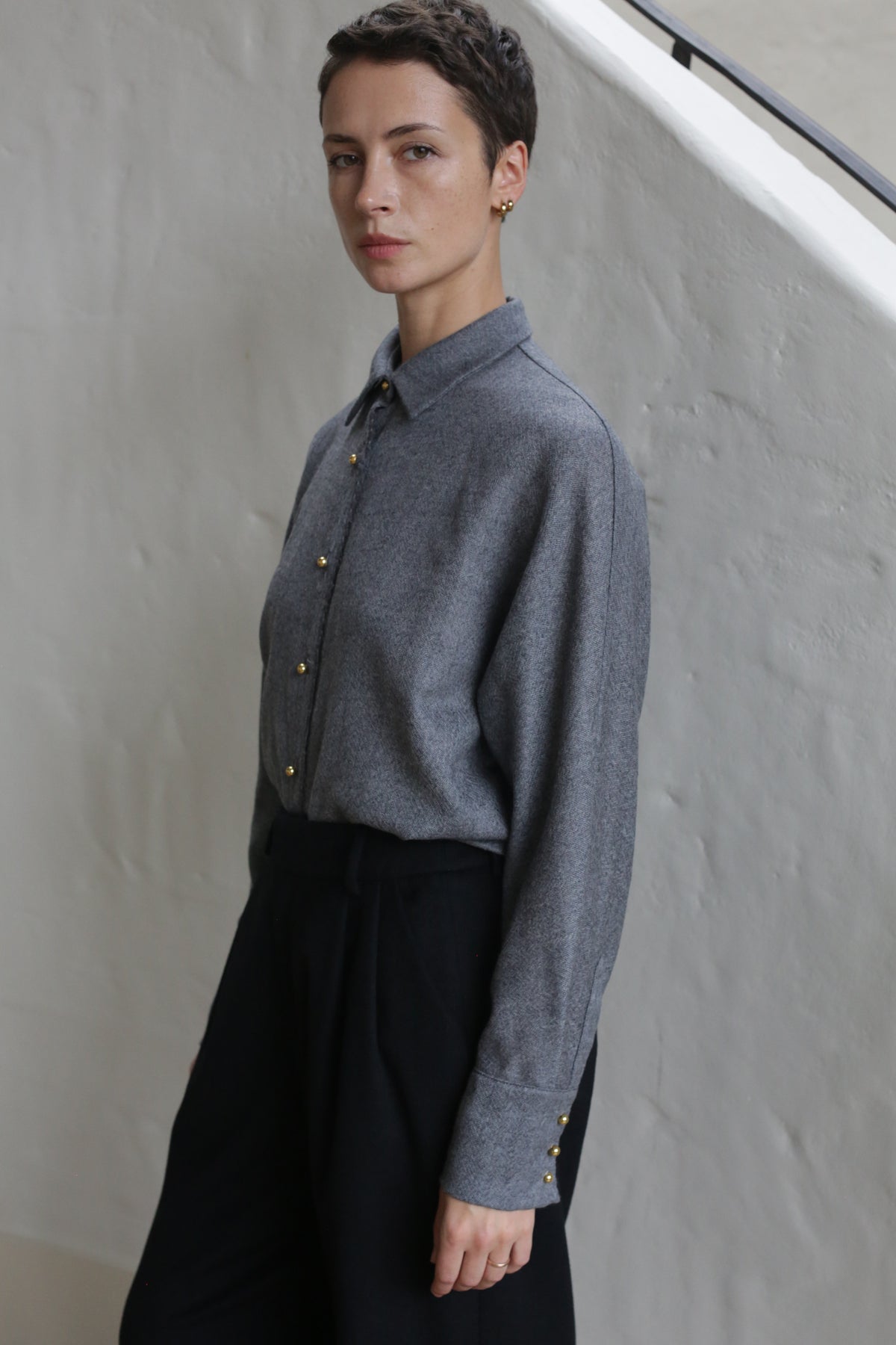 A person with short hair, dressed in the Work Shirt | Grey and black pants, stands beside a light-colored wall, looking calmly at the camera.