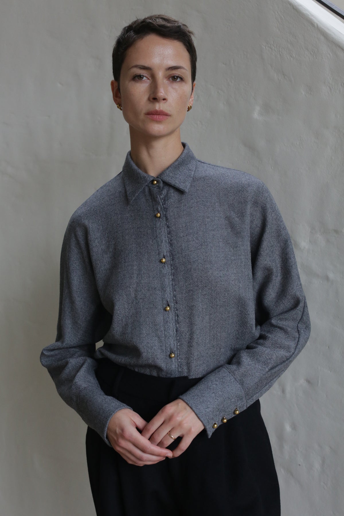 A person with short hair, wearing the Work Shirt | Grey and black pants, stands indoors against a light textured wall, looking directly at the camera with a neutral expression.