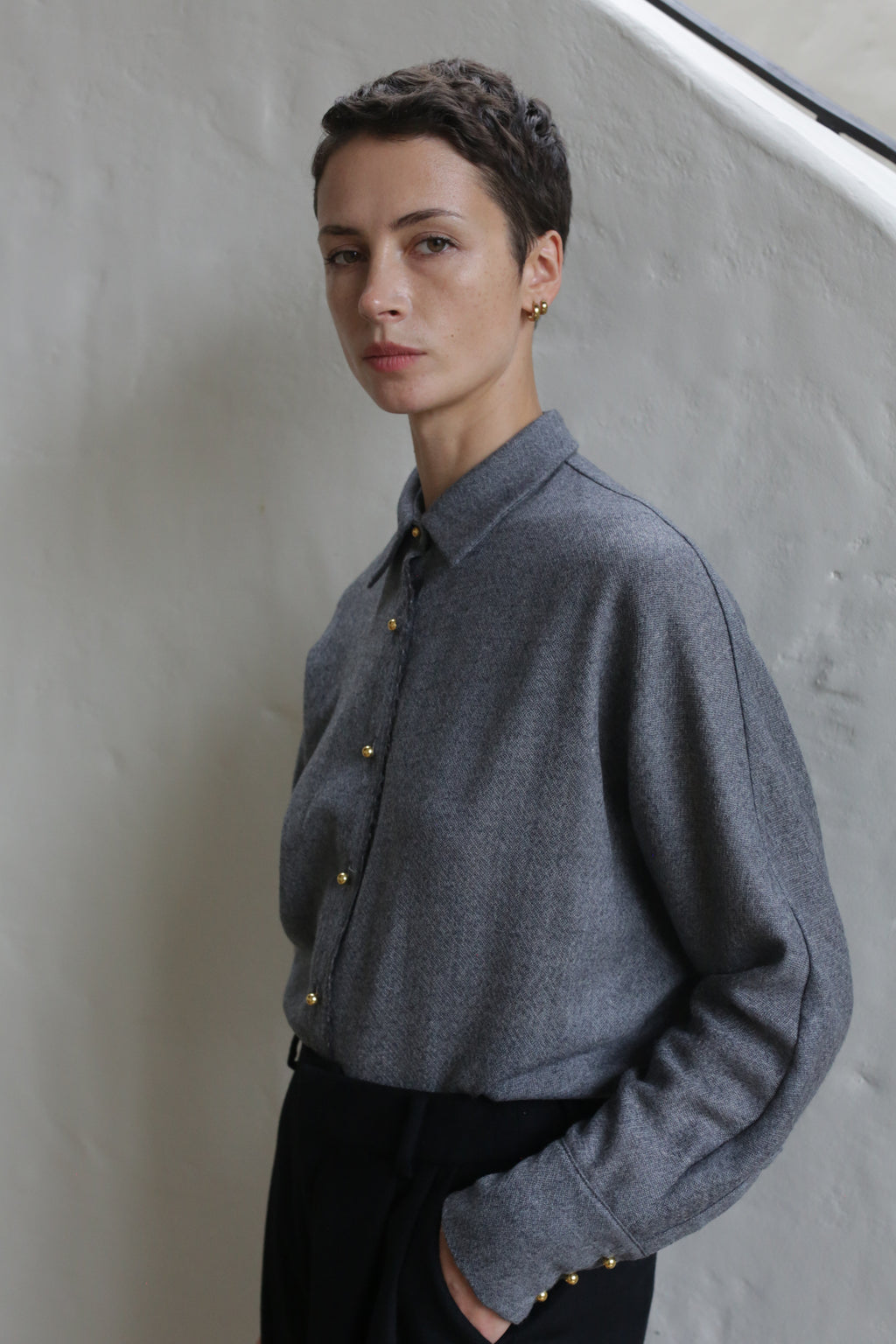 A person with short dark hair wears the Work Shirt | Grey and black pants, standing against a light textured wall, calmly looking at the camera with one hand in their pocket.