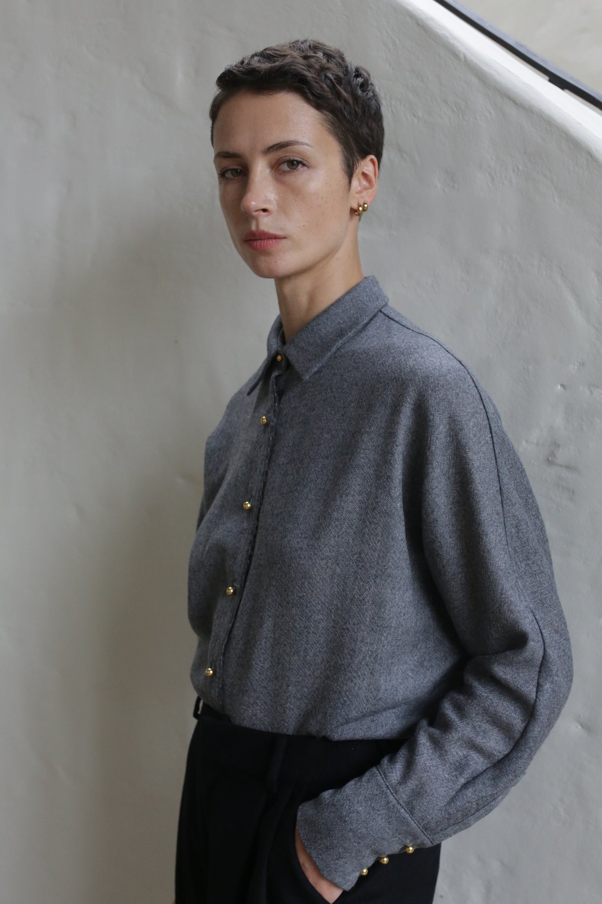 A person with short dark hair wears the Work Shirt | Grey and black pants, standing against a light textured wall, calmly looking at the camera with one hand in their pocket.