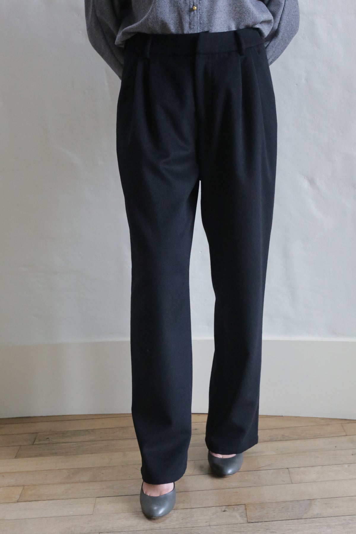 A person wearing dark Cashmere Pleated Trousers with a gray long-sleeve shirt tucked in and gray shoes stands on a wooden floor against a light-colored wall. Their upper face is not visible.