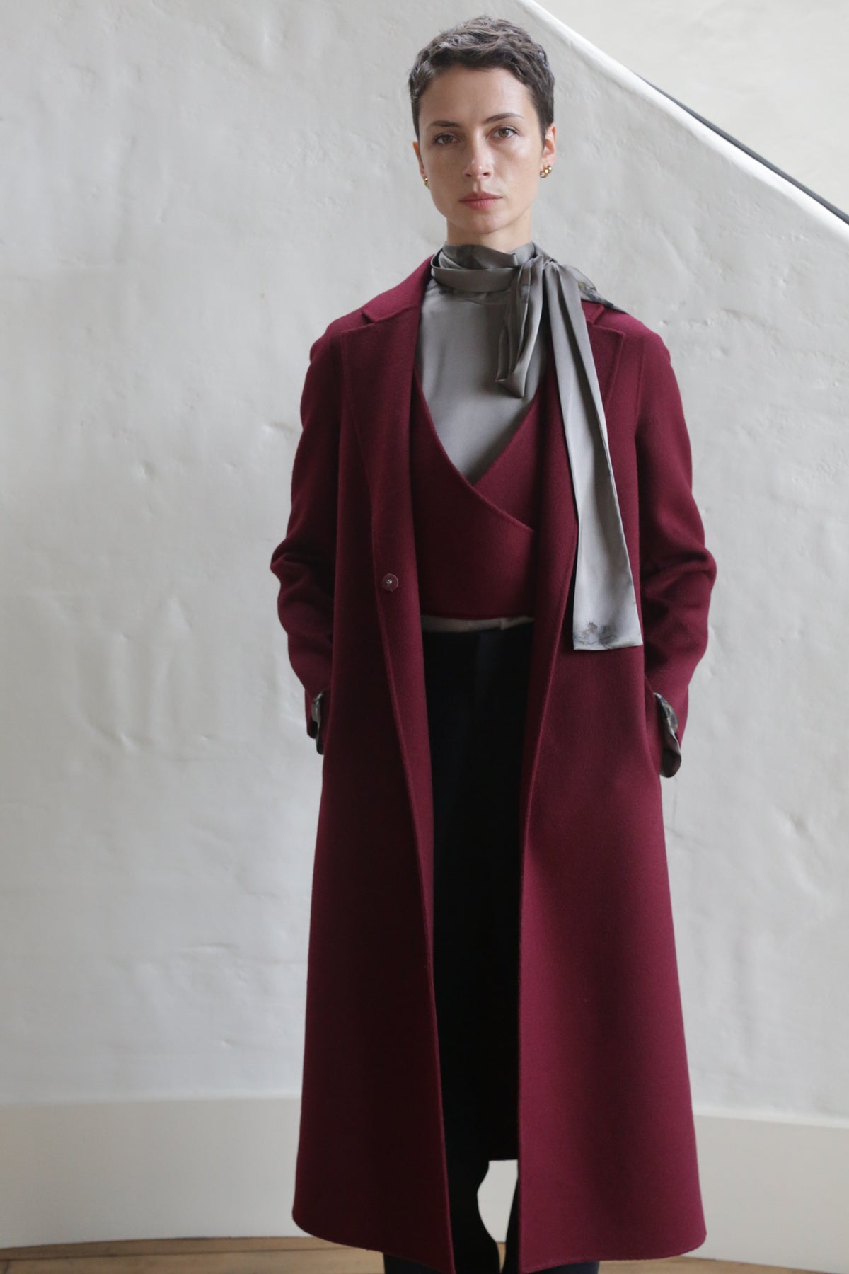 Indoors by a white textured wall and staircase, a person wears the X-Scarf Coat | Watermark Double Face Cashmere Burgundy over a gray top and scarf, hands in pockets.