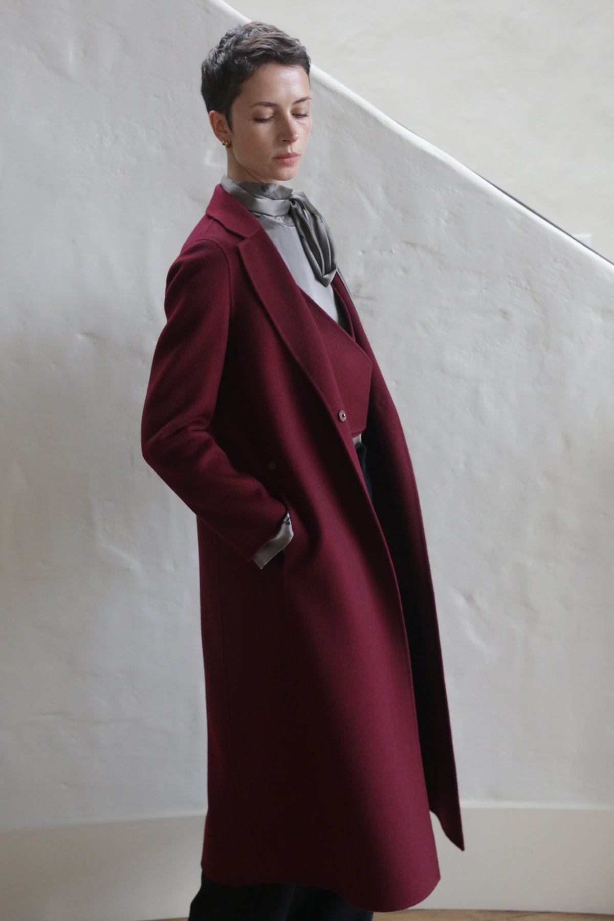 A person with short hair stands against a textured white wall, wearing the X-Scarf Coat | Watermark Double Face Cashmere Burgundy over a gray top, scarf loosely tied and one hand in their pocket, looking down thoughtfully.