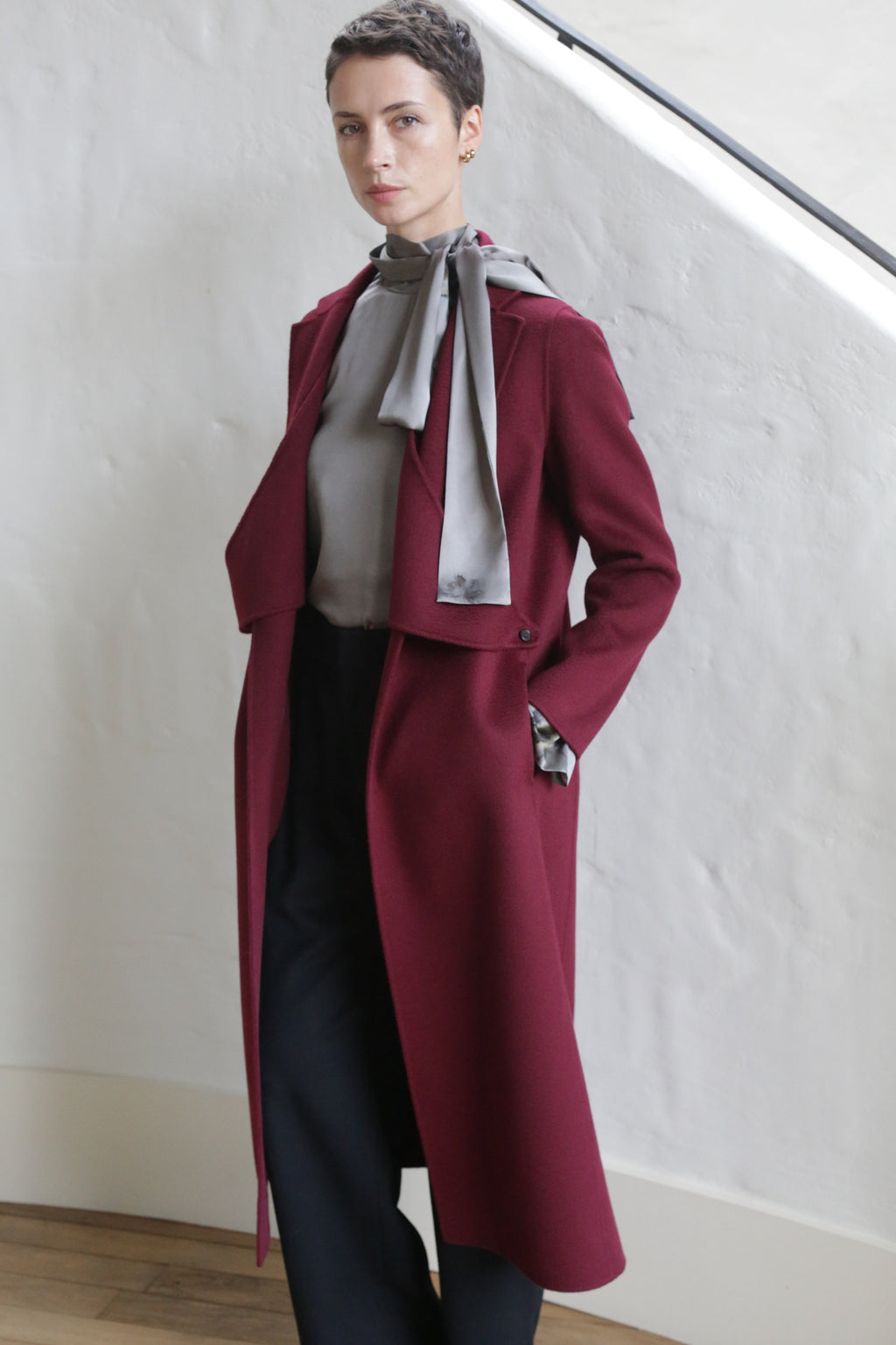 A person with short hair stands indoors, wearing the X-Scarf Coat | Watermark Double Face Cashmere Burgundy over a gray blouse and black pants, with one hand in their pocket and a neutral expression.