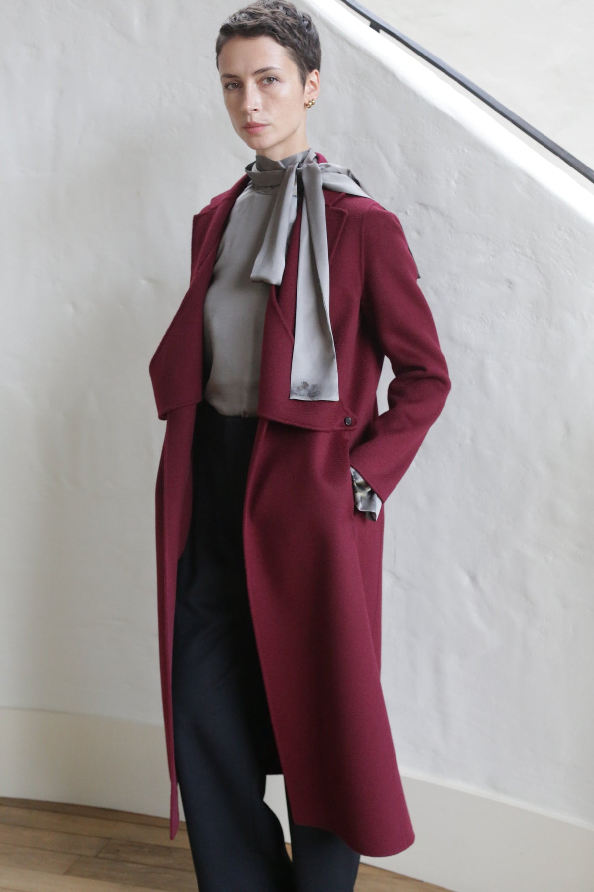 A person with short hair stands indoors, wearing the X-Scarf Coat | Watermark Double Face Cashmere Burgundy over a gray blouse and black pants, with one hand in their pocket and a neutral expression.
