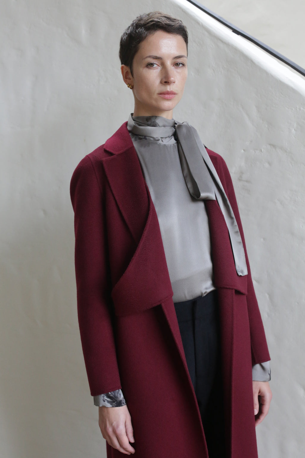 A person with short dark hair wears the X-Scarf Coat | Watermark Double Face Cashmere Burgundy, known for its innovative design, over a gray satin blouse with a tie at the neck, standing against a light-colored textured wall.