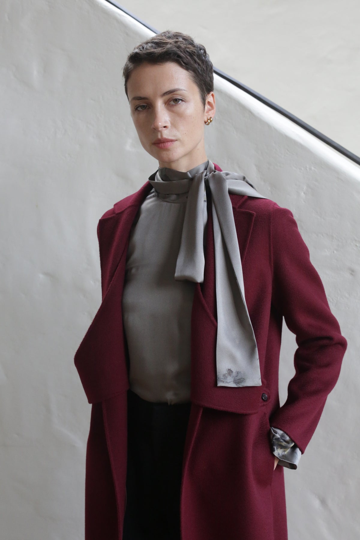 A person with short hair models the X-Scarf Coat | Watermark Double Face Cashmere Burgundy over a gray blouse, standing against a light wall and facing the camera with a neutral expression.