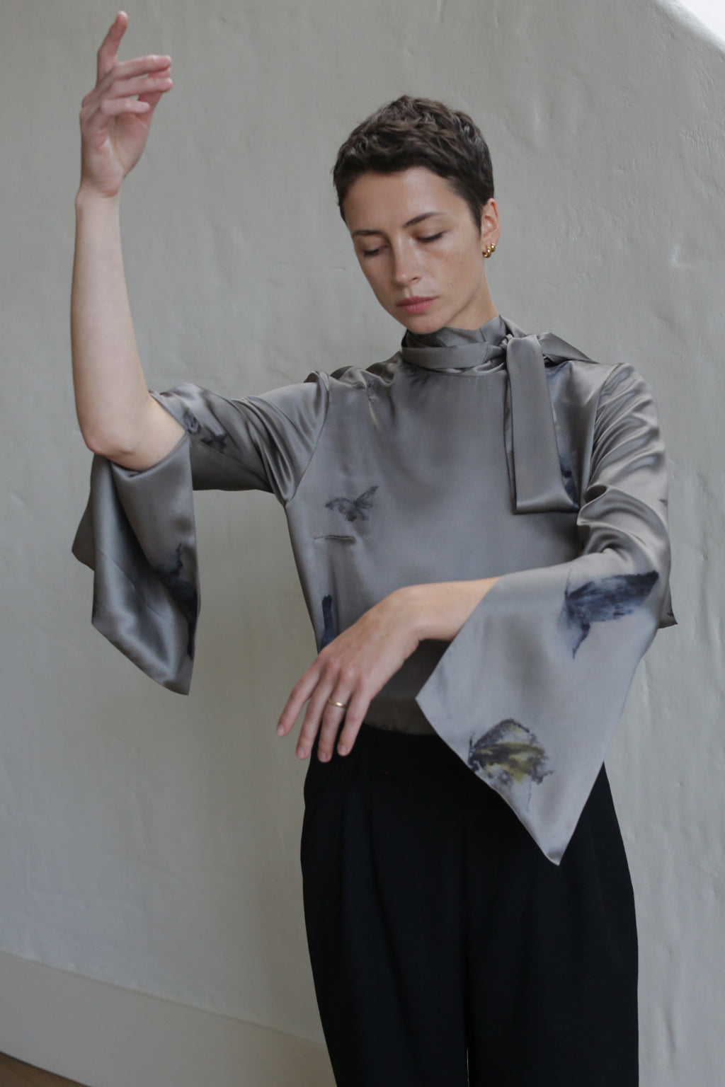 A person with short dark hair wears the Sash Blouse | Small Butterflies, featuring flared sleeves, paired with black pants, posing gracefully with one arm raised against a light-colored textured wall.