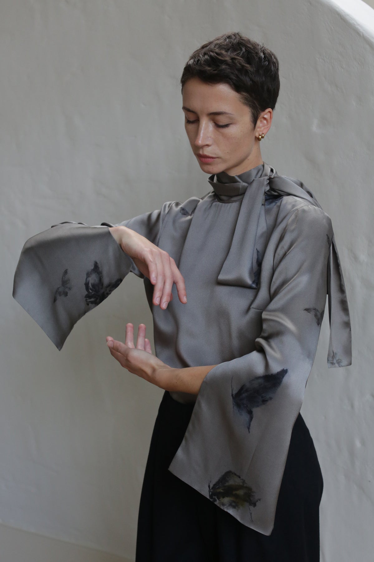 A person with short hair models the Sash Blouse | Small Butterflies, a gray, long-sleeved top with flared sleeves and a neck tie, posing gracefully with arms curved against a plain light background.