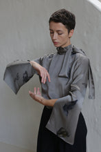 Load image into Gallery viewer, A person with short hair models the Sash Blouse | Small Butterflies, a gray, long-sleeved top with flared sleeves and a neck tie, posing gracefully with arms curved against a plain light background.