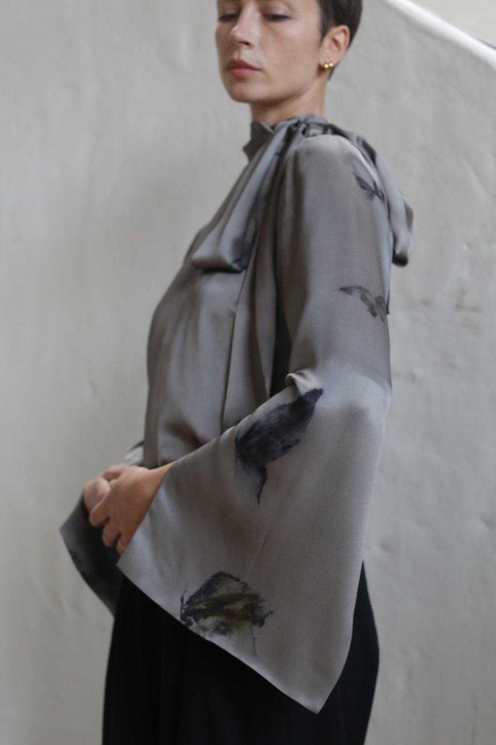 A person with short hair wears the Sash Blouse | Small Butterflies, a loose, silky gray top with wide sleeves and subtle dark floral patterns, standing against a plain light wall.