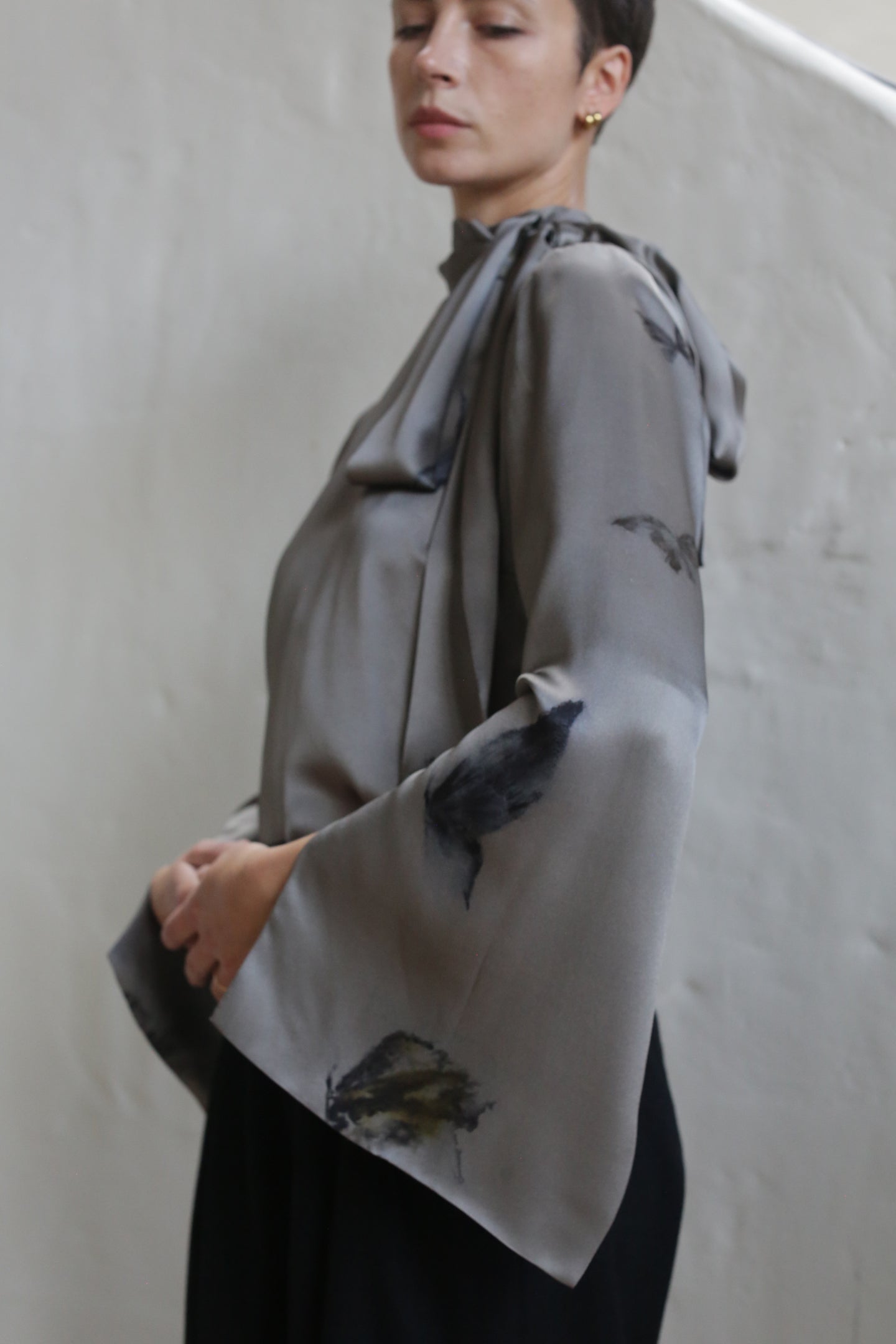 A person with short hair wears the Sash Blouse | Small Butterflies, a loose, silky gray top with wide sleeves and subtle dark floral patterns, standing against a plain light wall.